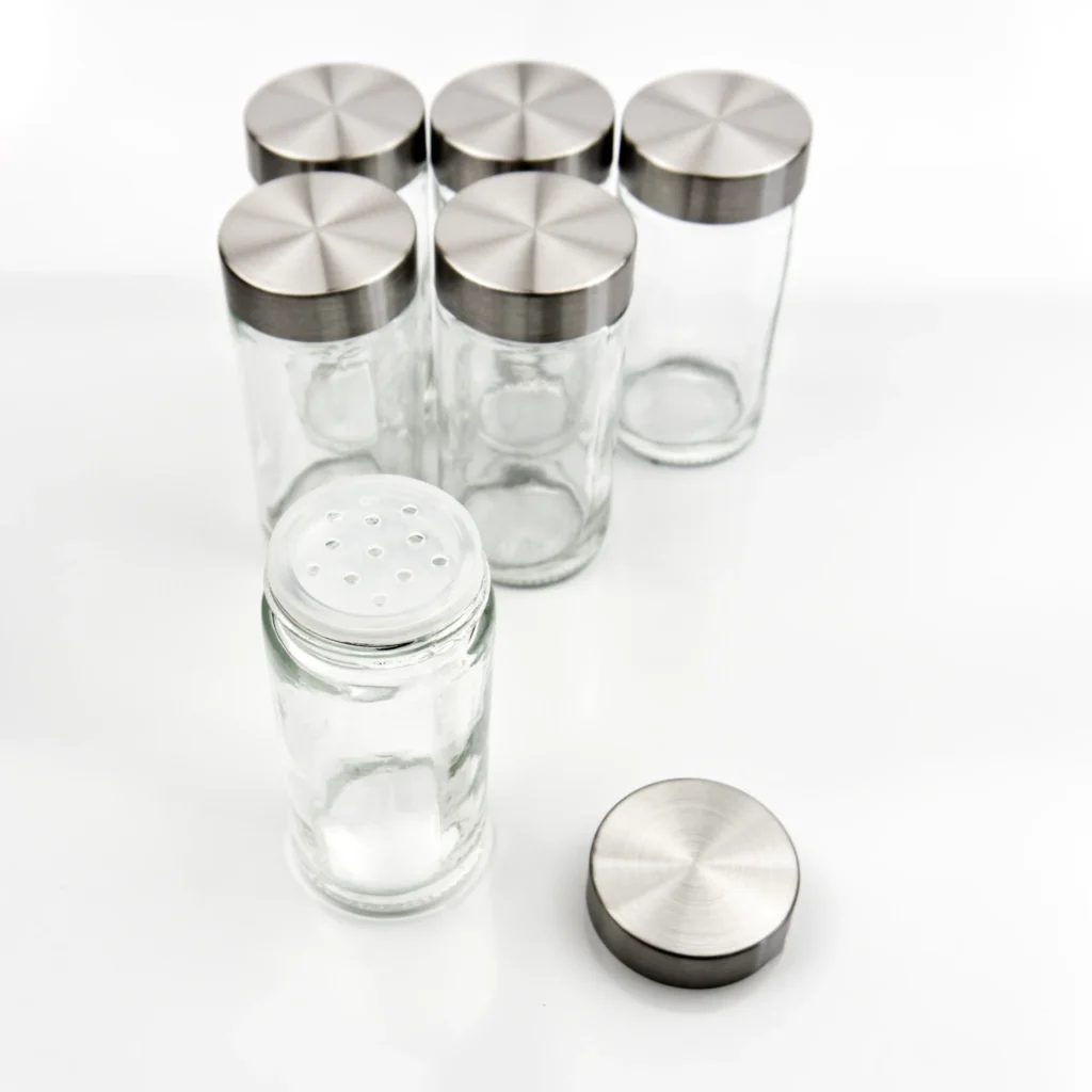 Stikla garšvielu burciņu komplekts ar sudraba metāla vākiem, priekšplānā viena burciņa ar caurumotu dozatora vāciņu.